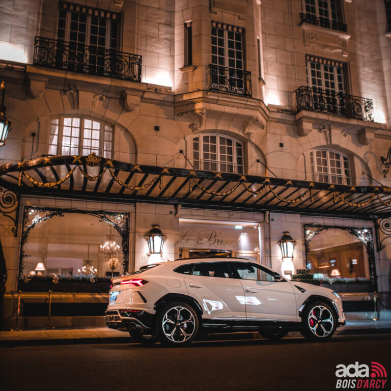 Lamborghini Urus blanc devant l'entrée illuminée d'un hôtel le soir, voiture de mariage à louer pour la Seine-Saint-Denis